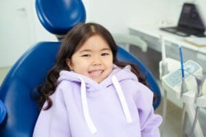 Girl in purple sweatshirt smiles in a dentist's chair for her pediatric dentistry appointment.